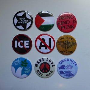 May include: A collection of nine circular pin-back buttons featuring various designs and text. The buttons display slogans including Rock Against Racism, Being Kind Is Punk, No ICE, No AI, Talk to Bugs Not Cops, Make Peace Not War, and Organize.