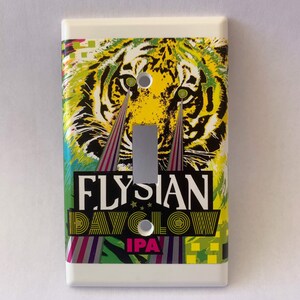 May include: A white light switch cover with a colorful graphic of a tiger's face with green eyes and the text "Flysian Dayglow IPA".