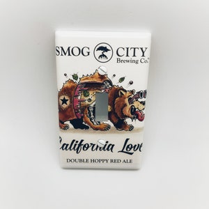 May include: A white light switch cover with a cartoon illustration of a bear wearing sunglasses and a red tongue sticking out. The bear is holding a beer bottle and the text "SMOG CITY Brewing Co." is at the top of the cover. The text "California Love" and "DOUBLE HOPPY RED ALE" are at the bottom of the cover.