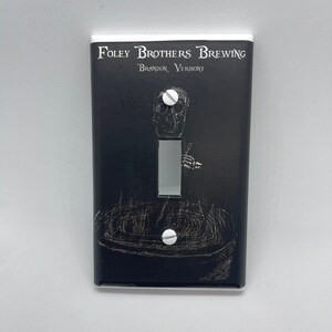 May include: Black light switch cover with a white border. The cover features a skull and crossbones graphic with the text "FOLEY BROTHERS BREWING" and "BRANDON, VERMONT".