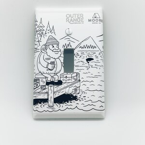 May include: A white light switch cover with a black and white illustration of a person fishing on a dock. The illustration includes text that says "Outer Range Brewing Co." and "Moore Denver, CO".