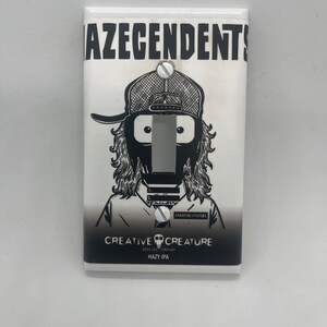 May include: A white light switch cover with a black and white illustration of a person wearing a hat and a mask. The text "AZEGENDENTS" is at the top of the cover. The text "CREATIVE CREATURE BREWING COMPANY HAZY IPA" is at the bottom of the cover.
