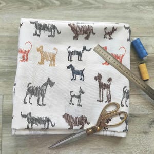 May include: White fabric with a repeating pattern of black, brown, and orange dogs. A pair of gold scissors, a ruler, and spools of thread are on a wooden surface.