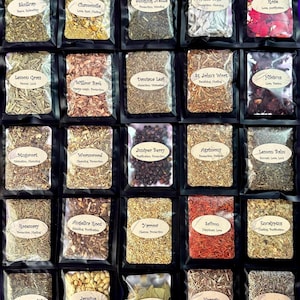 May include: A collection of twenty-five black resealable pouches containing various dried herbs, flowers, and botanicals. Each pouch features a white oval label with the name of the herb and its associated properties printed in black text.