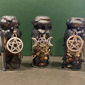 May include: Three glass bottles with black wax seals and silver charms. Each bottle contains herbs and stones. Charms include a pentacle, a triple moon, and another pentacle. The bottles are on a dark surface.