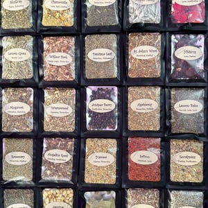 Puede incluir: Una colección de veinticinco pequeñas bolsas negras que contienen diversas hierbas y plantas secas. Cada bolsa cuenta con una ventana transparente y una etiqueta beige con el nombre de la hierba, como Skullcap, Chamomile, Stinging Nettle, White Sage y Rose.