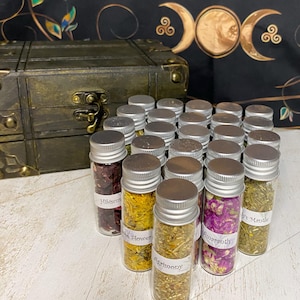 May include: A collection of small glass jars filled with dried herbs and flowers. The jars have silver lids and labels with names like "Hibiscus", "St. John's Flower", "Harmony", "Angelica", and "Lady's Mantle".