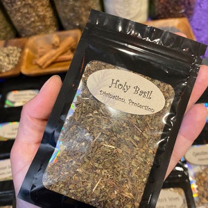 Puede incluir: Una bolsa negra resellable que contiene hojas secas de albahaca sagrada. El frente presenta una ventana transparente y una etiqueta ovalada de color canela con el texto Holy Basil, Divination, Protection en fuente marr&oacute;n.
