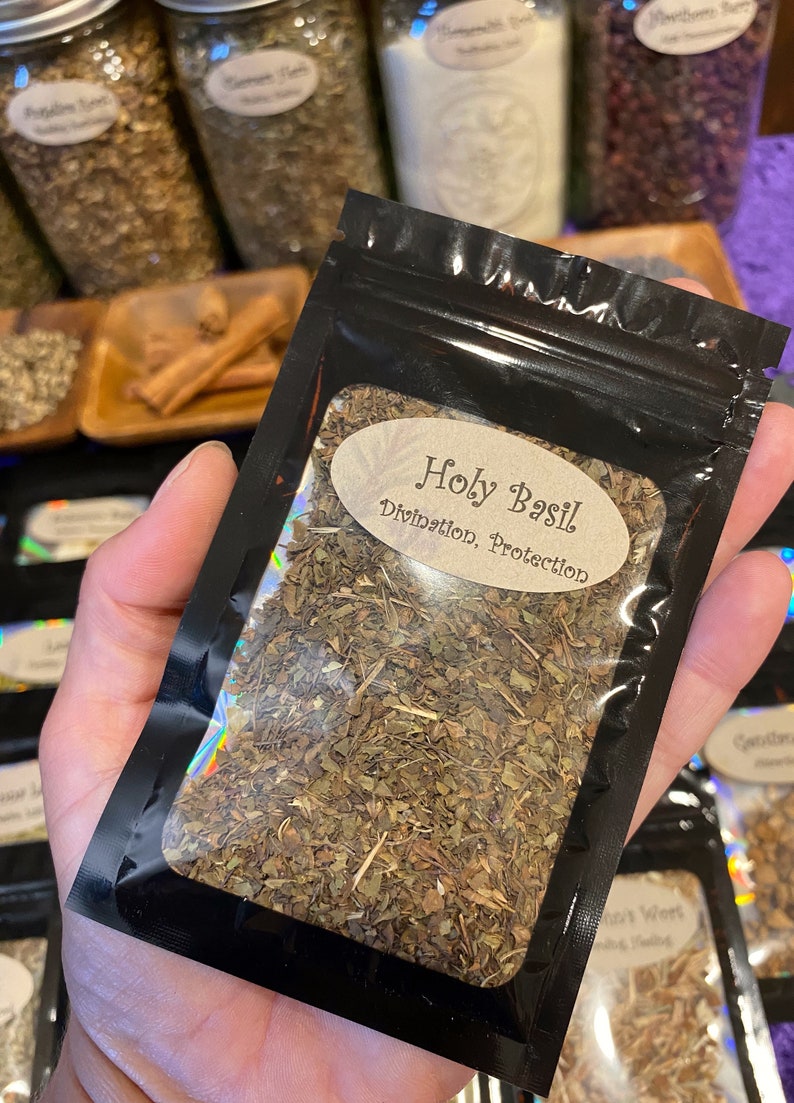 Puede incluir: Una bolsa negra resellable que contiene hojas secas de albahaca sagrada. El frente presenta una ventana transparente y una etiqueta ovalada de color canela con el texto Holy Basil, Divination, Protection en fuente marr&oacute;n.