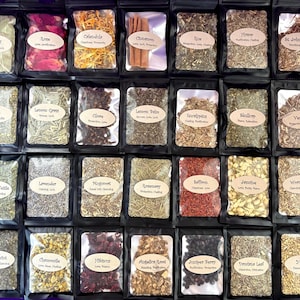Puede incluir: Una colecci&oacute;n de veintiocho bolsas negras que contienen diversas hierbas y productos bot&aacute;nicos secos. Cada bolsa cuenta con una ventana transparente y una etiqueta beige con el nombre de la hierba y sus propiedades, como laurel, rosa y canela.
