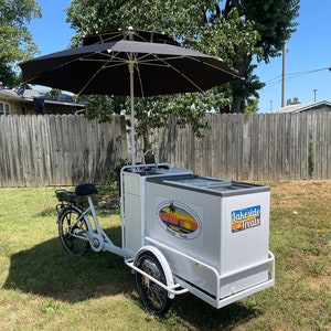 May include: White three-wheeled bicycle food cart with a black umbrella, a cooler, and a sign that says "Lakeside Treats".