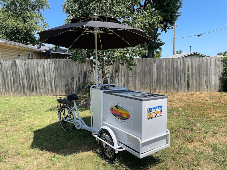 May include: White three-wheeled bicycle food cart with a black umbrella, a cooler, and a sign that says "Lakeside Treats".