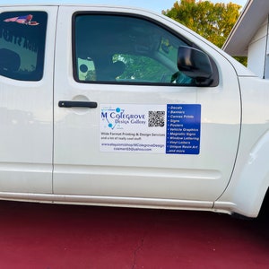 May include: A white pickup truck with a blue and white sign on the side that says "M Colegrove Design Gallery". The sign also includes a QR code and a list of services offered, including decals, banners, canvas prints, signs, posters, vehicle graphics, magnetic signs, window lettering, vinyl letters, unique resin art, and more.