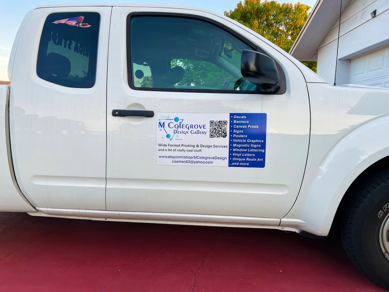 May include: A white pickup truck with a blue and white sign on the side that says "M Colegrove Design Gallery". The sign also includes a QR code and a list of services offered, including decals, banners, canvas prints, signs, posters, vehicle graphics, magnetic signs, window lettering, vinyl letters, unique resin art, and more.
