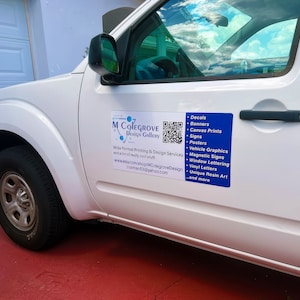 May include: White pickup truck with a blue and white sign on the side. The sign says "M Colegrove Design Gallery" and lists a variety of printing and design services, including decals, banners, canvas prints, signs, posters, vehicle graphics, magnetic signs, window lettering, vinyl letters, unique resin art, and more. A QR code is also included on the sign.