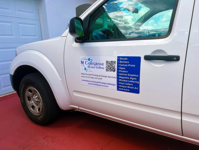 May include: White pickup truck with a blue and white sign on the side. The sign says "M Colegrove Design Gallery" and lists a variety of printing and design services, including decals, banners, canvas prints, signs, posters, vehicle graphics, magnetic signs, window lettering, vinyl letters, unique resin art, and more. A QR code is also included on the sign.