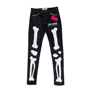 Può includere: Jeans in denim nero con design a ossa bianche. I jeans presentano dettagli strappati e una toppa con la scritta "THE SKINS COLLECTION". Un'etichetta rossa pende dal passante. Un capo di moda unico.