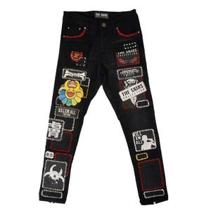 May include: Black denim jeans with multiple patches featuring various designs and text. Patches include a colorful flower, a skull, and the text "KILL EM ALL". Red stitching accents the pockets and seams.