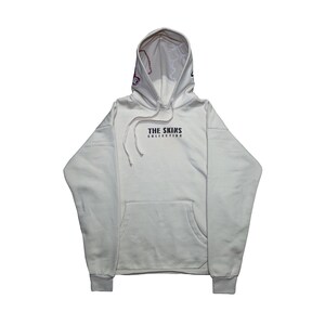 May include: A cream-coloured hoodie with a front pocket and drawstring hood. The text "THE SKINS COLLECTION" is printed in black on the chest. The hood has embroidered details. The cuffs and hem are ribbed.