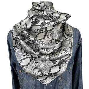 May include: A square scarf featuring a monochromatic snakeskin pattern in shades of grey, black, and white. The scarf is tied in a knot at the neck and draped over a blue denim shirt. The fabric has a smooth, slightly reflective finish throughout.