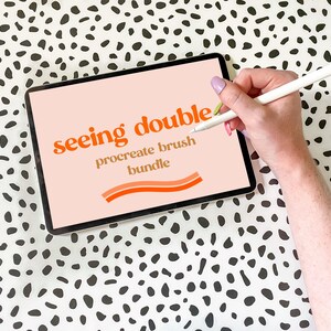 May include: A digital tablet displaying a pink background with the text "seeing double procreate brush bundle" in orange and yellow text. A hand is holding a white stylus and drawing on the screen.
