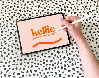 May include: A digital drawing tablet with a pink background and the text "kellie procreate brush" in orange. A hand is holding a white stylus and drawing a curved line on the screen.