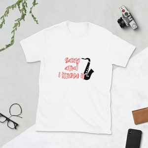 Puede incluir: Camiseta blanca con un gráfico de saxofón negro y el texto "Sax and I know it" en rojo.