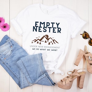 May include: White t-shirt with the text "Empty Nester Under New Management We Do What We Want" and a graphic of mountains and trees. The shirt is paired with blue jeans, brown sandals, and sunglasses.