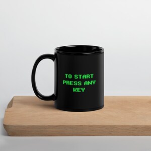 Puede incluir: Taza de cerámica negra con un gráfico de texto verde que dice "TO START PRESS ANY KEY".