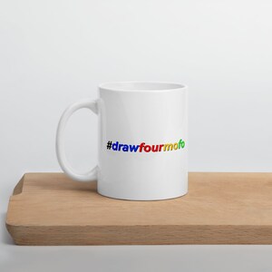 May include: A white ceramic mug with a curved handle. The mug features the colorful text "#drawfourmofo" in blue, red, yellow, and green. The mug is placed on a wooden surface against a white background.