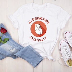 Puede incluir: Camiseta blanca con un círculo rojo y un gráfico de corazón blanco. El texto "All bleeding stops... eventually" está impreso en la camiseta.