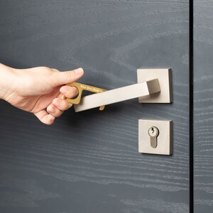 Może przedstawiać: Złoty przyrząd do klamki z napisem "Wash Your Damn Hands" służy do otwierania srebrnej klamki. Drzwi i klamka zamontowane są na szarych drewnianych drzwiach.