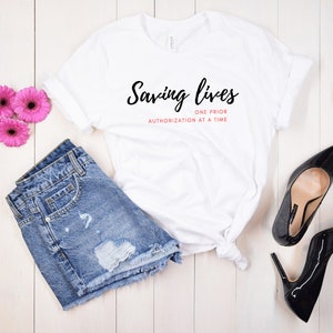 Può includere: Maglietta bianca con il testo "Saving lives one prior authorization at a time" in lettere nere e rosa. La maglietta è appoggiata su una superficie di legno bianco con un paio di pantaloncini di jeans blu, due fiori rosa e una bottiglia di champagne.