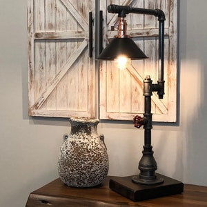 May include: A black pipe table lamp with a copper accent and a black lampshade. The lamp is on a wooden table next to a white ceramic vase.