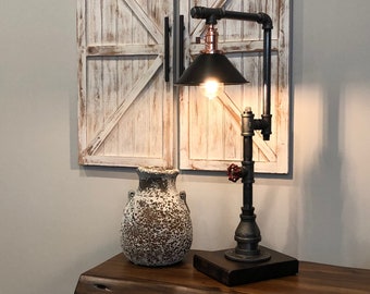 May include: A black pipe table lamp with a copper accent and a black lampshade. The lamp is on a wooden table next to a white ceramic vase.