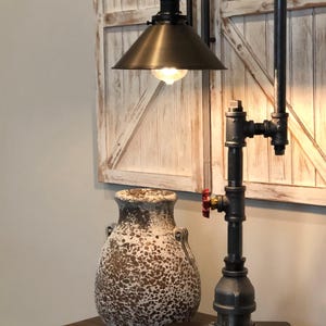 May include: Industrial-style table lamp with a dark metal pipe frame, a bronze-colored lampshade, and a visible lightbulb. The lamp features a red valve detail and sits on a dark wooden base. A decorative vase with a speckled pattern is next to the lamp.