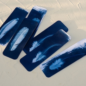 May include: Five blue rectangular bookmarks with white feather images printed on them. The bookmarks are arranged in a fan shape on a white surface.
