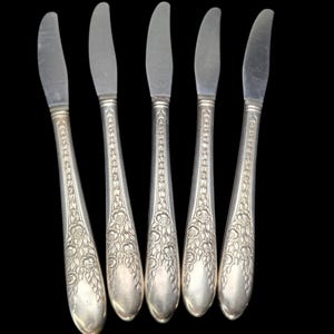 Puede incluir: Cinco cuchillos de mantequilla plateados con diseños florales y de hojas en los mangos. Las hojas son lisas y curvas. Los mangos tienen un patrón detallado de rosas y hojas. Los cuchillos están dispuestos sobre un fondo negro.