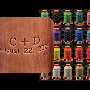 May include: A brown leather journal with the text "C+D August 22, 2021" embossed on the front. The journal is surrounded by a variety of colored thread spools.