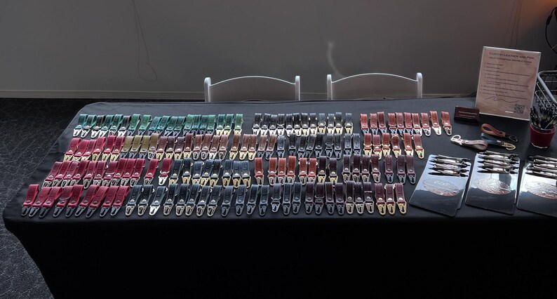 May include: A display of leather belts in various colors, including red, green, black, and brown, with silver-colored buckles. The belts are arranged on a black table, showcasing a wide selection of styles and hues.