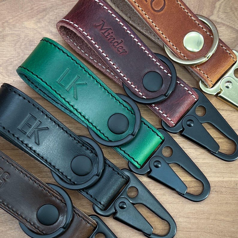 May include: Close-up of several leather keychains in various colors, including black, green, and brown. Each keychain features a metal clasp and a leather strap with embossed initials or a name. The keychains are arranged on a wooden surface.