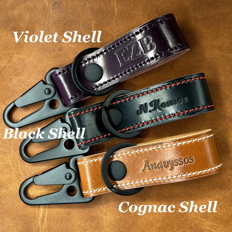 May include: Three leather keychains in violet, black, and cognac colors. Each keychain features a metal clasp and a ring. The violet keychain has the initials "EZB", the black keychain has "N. Kosmos", and the cognac keychain has "Andvyssos".