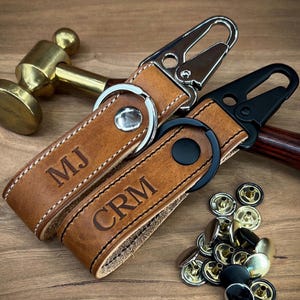May include: Two personalized leather keychains with silver and black hardware. The brown leather key fobs are embossed with initials "MJ" and "CRM". White stitching accents the edges. A collection of metal snaps is visible.