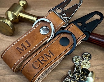 May include: Two personalized leather keychains with silver and black hardware. The brown leather key fobs are embossed with initials "MJ" and "CRM". White stitching accents the edges. A collection of metal snaps is visible.