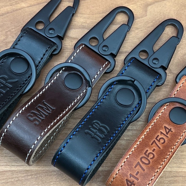 Four 1” wide and 5” long American Horween leather keychains with black metal sling lever snaps and matching key rings. The keychains are brown, black, and tan. The keychains are handmade to order in Houston, Texas by CustomLeatherAndPen. The brown keychain has the initials 'SMM' engraved on it. The black keychain has the initials 'JJB' engraved on it. The tan keychain has the numbers '41-705-7514' engraved on it. The fourth keychain is black and has no visible engraving.