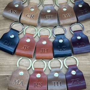 May include: A collection of 12 leather keychains with metal rings. Each keychain has a different two-letter monogram in a silver font. The keychains are in various colors, including brown, black, burgundy, and tan.