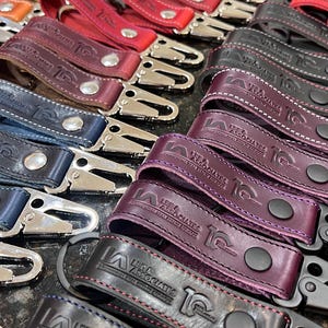 May include: A collection of leather keychains in various colors, including red, brown, and purple. Each keychain features a metal clasp and the embossed text "LEE & ASSOCIATES". The keychains are arranged in neat rows, showcasing the variety of colors and designs.