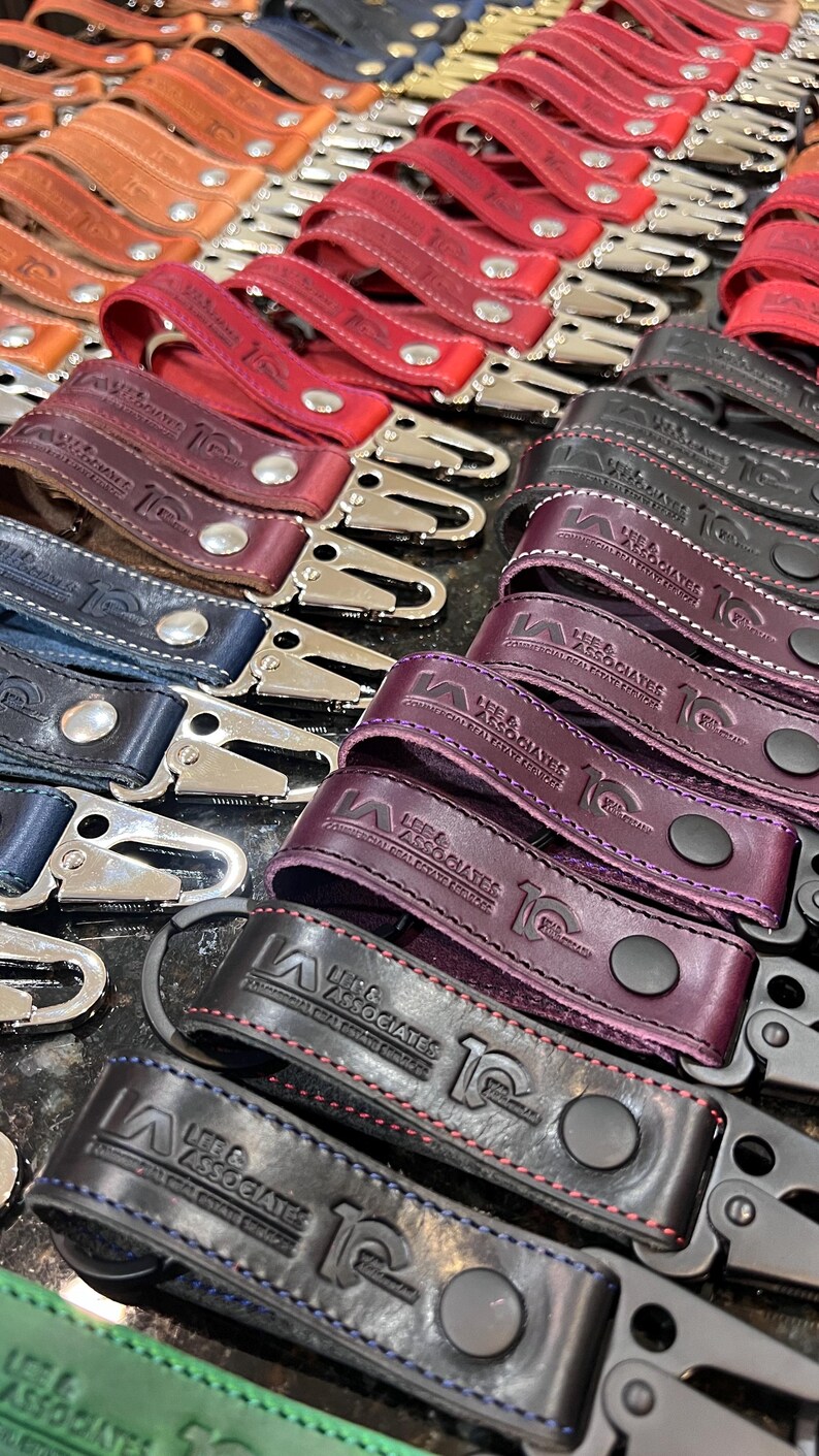 May include: A collection of leather keychains in various colors, including red, brown, and purple. Each keychain features a metal clasp and the embossed text "LEE & ASSOCIATES". The keychains are arranged in neat rows, showcasing the variety of colors and designs.