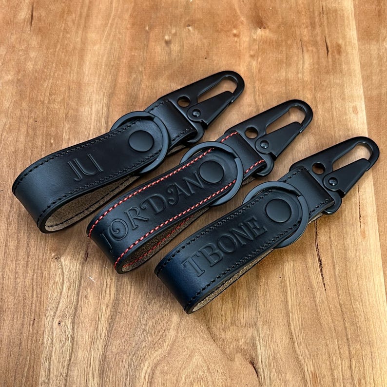 May include: Three black leather keychains with metal clasps are displayed on a wooden surface. Each keychain has a different name embossed on it: "JUL", "JORDAN", and "TBONE". The "JORDAN" keychain has red stitching.