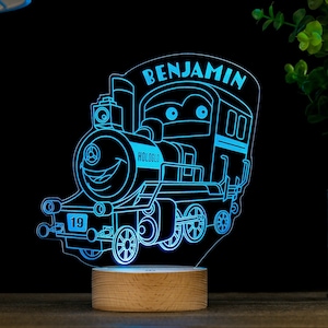 May include: A blue LED night light shaped like a cartoon train with the name "BENJAMIN" on the side. The train has a smiling face and the number "19" on the front. The light is sitting on a wooden base.